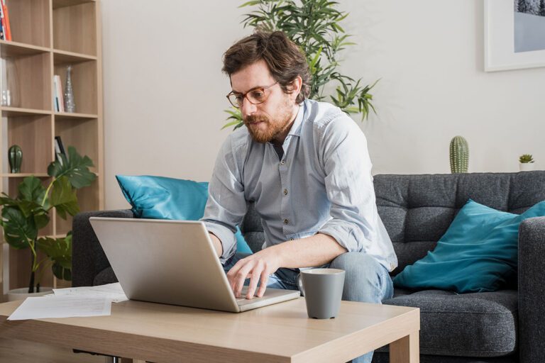 Ein Mann mit Brille sitzt auf einem grauen Sofa und arbeitet in seinem Homeoffice an einem Laptop, an einem hölzernen Couchtisch mit einer Tasse und Papieren, in einem Wohnzimmer mit Pflanzen und blauen Polstern. IFF Institut für Fach- und Führungskräfte
