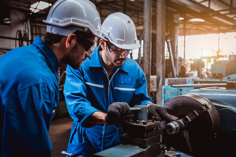 Zwei Arbeiter in blauen Overalls und weißen Schutzhelmen bedienen Industriemaschinen in einer Fabrik. Beide, möglicherweise ein Zerspanungsmechaniker und ein Industriemeister Metall, tragen Schutzbrillen; der eine stellt die Maschine ein, während der andere sie genau beobachtet. IFF Institut für Fach- und Führungskräfte