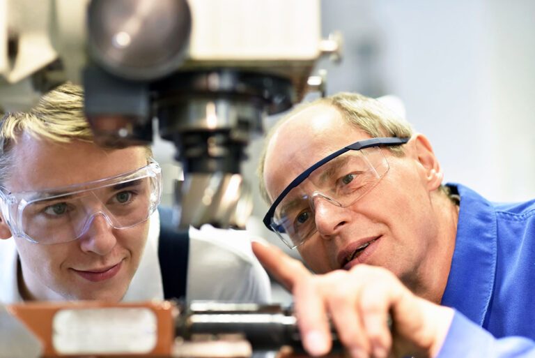 Zwei Männer mit Schutzbrillen untersuchen und besprechen eine Maschine und verdeutlichen damit die Bedeutung des Handwerks in einer Werkstatt - vielleicht sogar in einem Moment der Unternehmensnachfolge. IFF Institut für Fach- und Führungskräfte